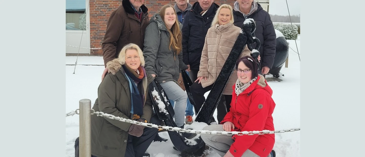 Fehnker Spaoßmaokers_1 (1920x1080).jpg Acht Personen posieren lächelnd im Schnee vor einem Anker beim Dorfgemeinschaftshaus Wilhelmsfehn.