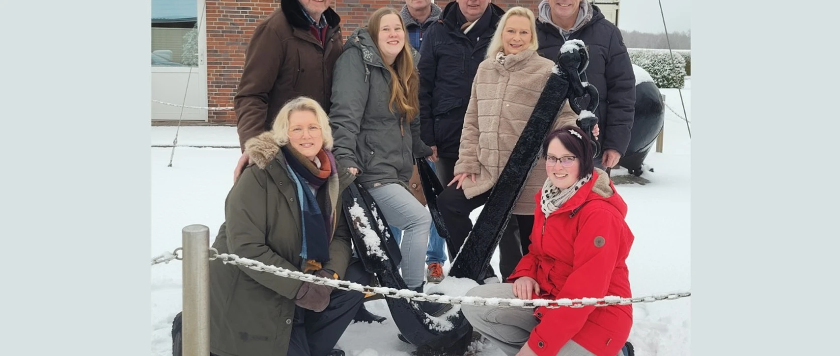 Fehnker Spaoßmaokers_1 (1920x1080).jpg Acht Personen posieren lächelnd im Schnee vor einem Anker beim Dorfgemeinschaftshaus Wilhelmsfehn.