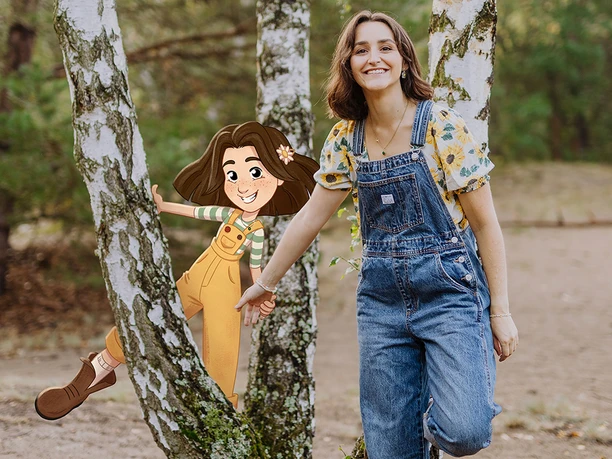 Lottis Welt in Büsum Eine Frau in Latzhose vor einem Baum. Sie hält ein Mädchen in der Hand, das aussieht wie eine Illustration. Diese trägt eine gelbe Latzhose und hat braune Haare.