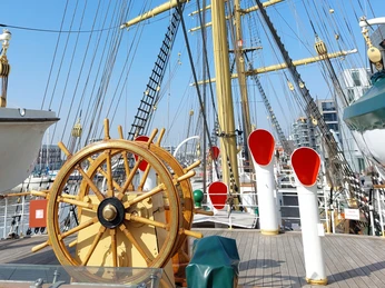 Schulschiff Deutschland 20220322_130222 (c) Isabel Wienberg_Erlebnis Bremerhaven_Ausschnitt.jpg Holzsteuerrad auf dem Deck des Schulschiffs Deutschland vor gelben Masten und Tauwerk im Hafen.