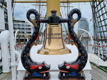 Glocke SSD (c) Hans L. Johnen_DSV.jpg Messingglöcke des Schulschiffs Deutschland zwischen geschnitzten, bunt bemalten Delfinen.