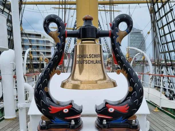 Glocke SSD (c) Hans L. Johnen_DSV.jpg Messingglöcke des Schulschiffs Deutschland zwischen geschnitzten, bunt bemalten Delfinen.