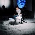 RETINA MANEUVER Person kniet in dichter Nebelwolke vor Technikgeräten, im Hintergrund leuchtet ein blaues Projektionbild.Person kneeling in a dense cloud of fog in front of technical equipment, a blue projection image glows in the background.