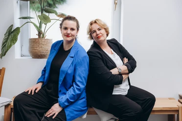 zwei Damen sitzen auf einer Bank vor einer weißen Wand