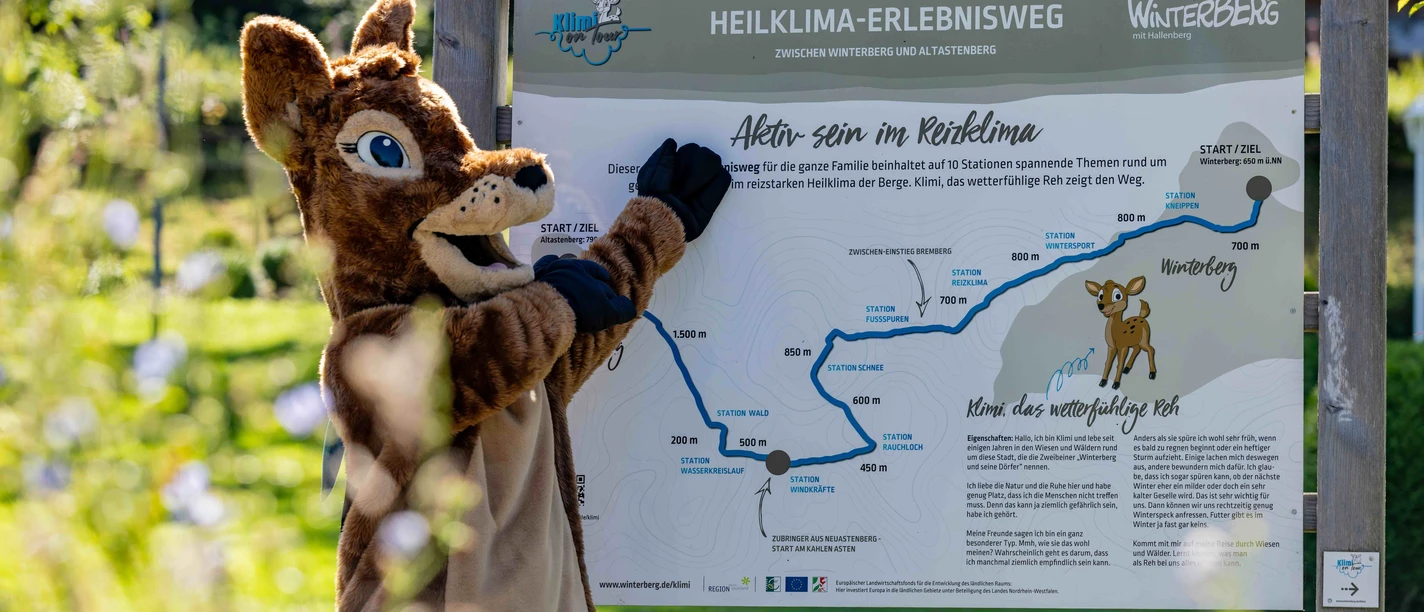 Waldi zeigt auf das Heiklima-Erlebnisweg-Schild
