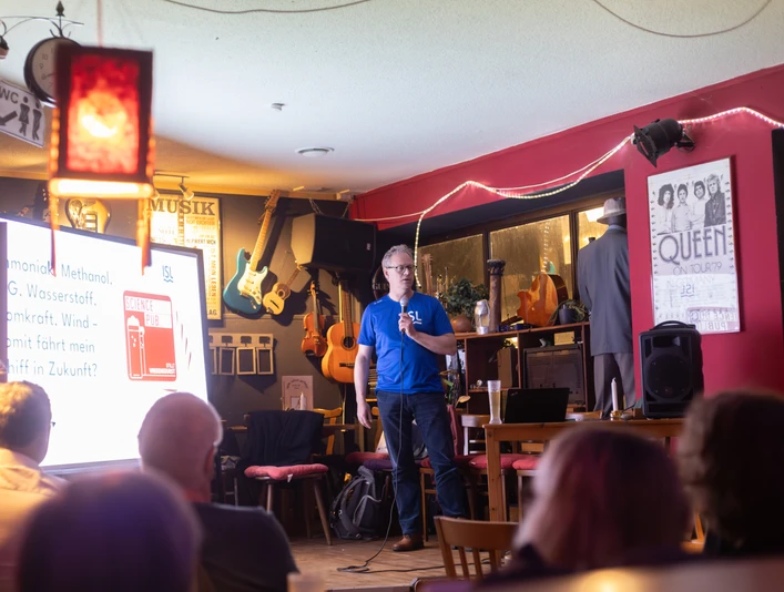 Meyer-Larsen_SGP25_(c)SebastianKühl.jpg Ein Mann hält in einer Bar einen Vortrag über Wasserstoff, Publikum hört aufmerksam zu.