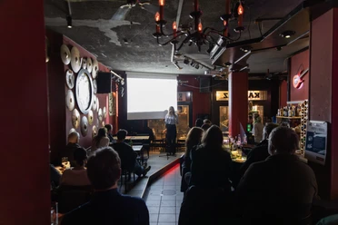 Menschen hören in einer Bar einer Vortragenden zu, die vor einer Leinwand auf der Bühne steht.