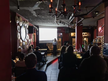 01_Kapovaz-13.jpg Menschen hören in einer Bar einer Vortragenden zu, die vor einer Leinwand auf der Bühne steht.