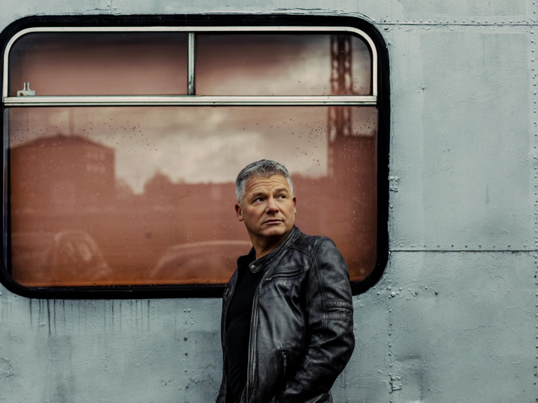 Andreas Winkelmann Mann in dunkler Lederjacke steht vor metallischer Wand mit Fenster und blickt nach rechts.