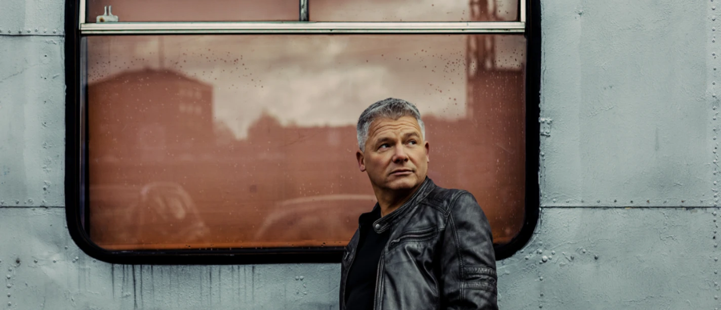 Andreas Winkelmann Mann in dunkler Lederjacke steht vor metallischer Wand mit Fenster und blickt nach rechts.