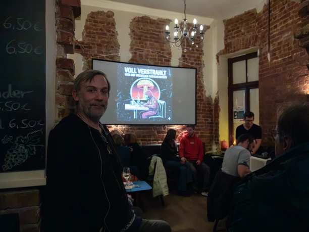 Walter_Geibert_SGP25_(c)SebastianKühl.jpg Mann in Bar mit Backsteinwänden, im Hintergrund Publikum und Leinwand mit Vortragstitel.