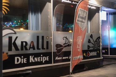 Eingangsbereich der Kneipe Kralli in Bremerhaven mit beleuchteten Fenstern und Werbefahne bei Nacht.