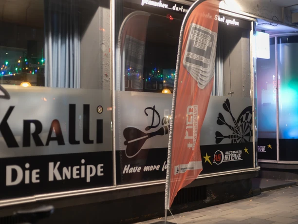 05_Kralli-15.jpg Eingangsbereich der Kneipe Kralli in Bremerhaven mit beleuchteten Fenstern und Werbefahne bei Nacht.