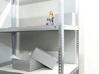 HisMus_(c)AstridOttens.jpg Eine Puppe im Rollstuhl steht auf einem Metallregal neben grauen Kartons in einem hellen Raum.