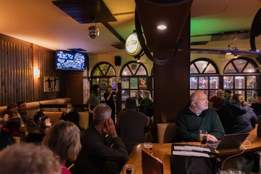 Menschen sitzen in einer Bar und hören einem Vortrag zu, während Getränke auf den Tischen stehen.