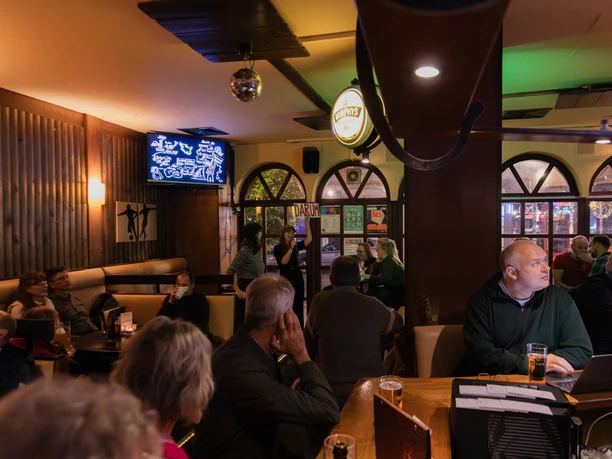 01_Rüssel-1.jpg Menschen sitzen in einer Bar und hören einem Vortrag zu, während Getränke auf den Tischen stehen.