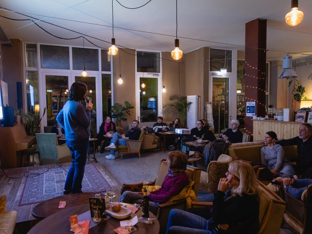 01_Findus-14.jpg Person hält Vortrag vor aufmerksamem Publikum in einem Café mit warmem Licht und entspannter Atmosphäre.