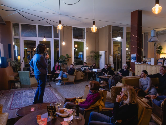 01_Findus-14.jpg Person hält Vortrag vor aufmerksamem Publikum in einem Café mit warmem Licht und entspannter Atmosphäre.
