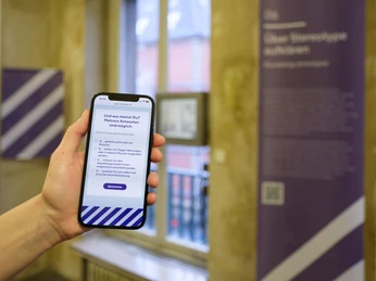 Intervention! Wie erzählen wir NS-Geschichte heute? Hand hält ein Smartphone mit Umfrageansicht in einem Ausstellungsraum des Museums.Hand holds a smartphone with a survey view in an exhibition room of the museum.