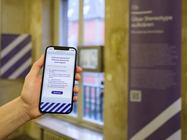 Intervention! Wie erzählen wir NS-Geschichte heute? Hand hält ein Smartphone mit Umfrageansicht in einem Ausstellungsraum des Museums.