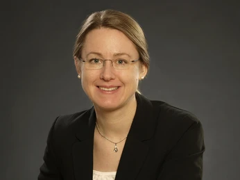 Julia Kahleyß_Fotograf-StephanGawlik.jpg Frau mit Brille und Hochsteckfrisur in dunklem Blazer vor neutralem Hintergrund, freundlich lächelnd
