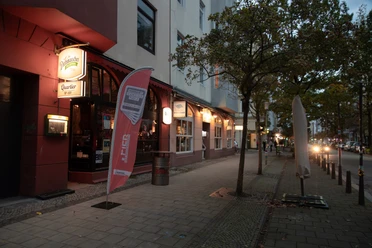 Beleuchteter Eingang des Quartier No. 159 an einer Abendstraße mit Bäumen und wenigen Passanten