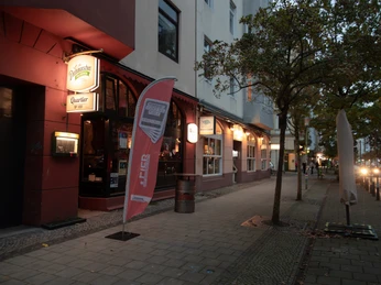 01_Q159-2.jpg Beleuchteter Eingang des Quartier No. 159 an einer Abendstraße mit Bäumen und wenigen Passanten