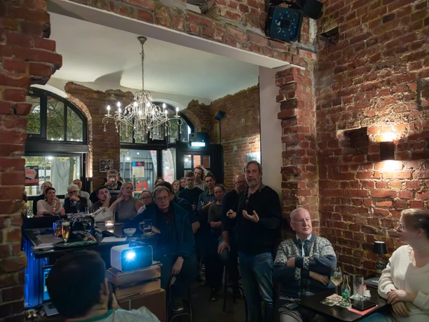 01_Q159-16.jpg Menschen verfolgen in rustikaler Bar mit Backsteinwänden einen Vortrag mit Projektion und Getränken.