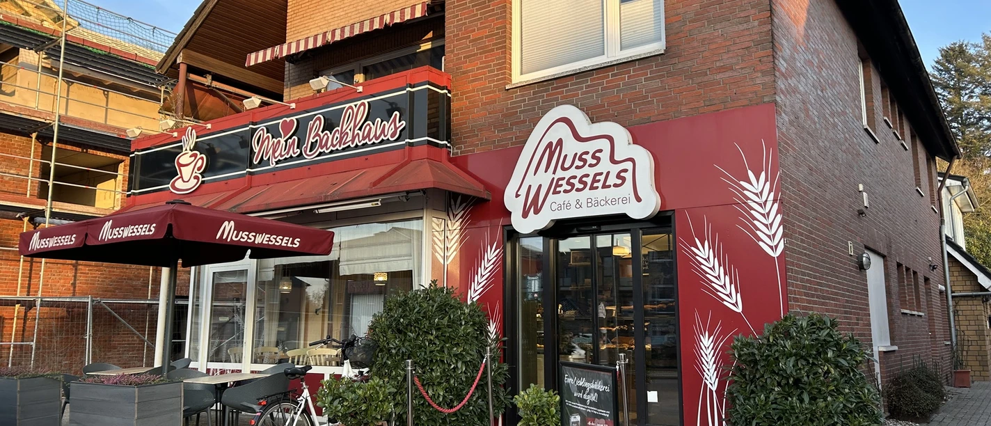baeckerei-musswessels-leer-loga.jpg Backsteinhaus mit rotem Fassadenbereich, Eingang zur Bäckerei Musswessels mit Terrasse.