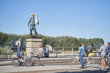 Leda - Markus Lüpertz Skulptur Leda von Markus Lüpertz: Frau mit Gans auf einem Sockel, Menschen mit Fahrrädern im Umfeld.