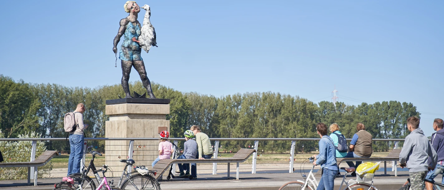 Leda - Markus Lüpertz Skulptur Leda von Markus Lüpertz: Frau mit Gans auf einem Sockel, Menschen mit Fahrrädern im Umfeld.