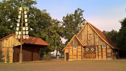 Historisches Bauernhaus mit Fachwerk, Backstein, Giebeln und freiem Hof, links ein Wappenpfahl.