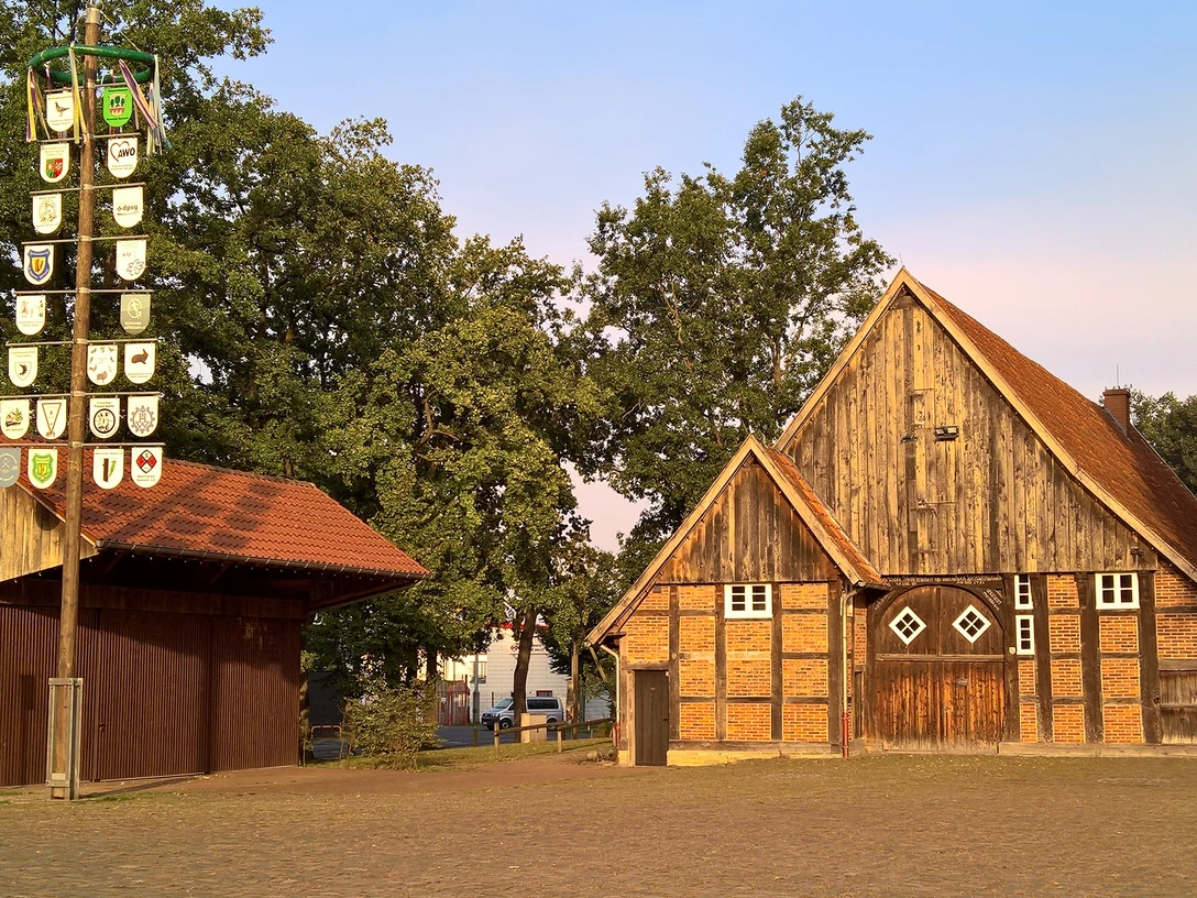 Historisches Bauernhaus mit Fachwerk, Backstein, Giebeln und freiem Hof, links ein Wappenpfahl.
