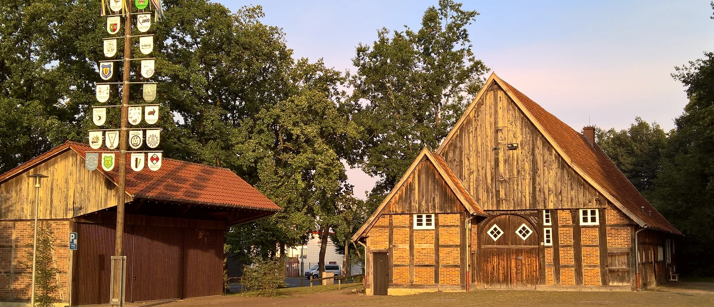Historisches Bauernhaus mit Fachwerk, Backstein, Giebeln und freiem Hof, links ein Wappenpfahl.