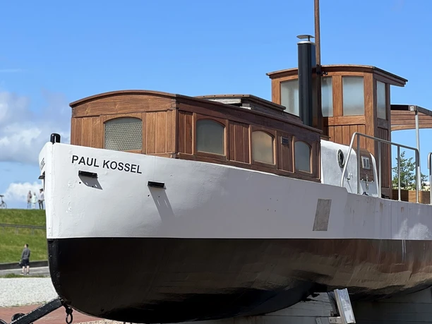 Paul Kossel 220522 InstaWalk Vom Ursprung 27 (c) Sandra Braumann_Erlebnis Bremerhaven_WEB.jpg Historisches Betonrumpfschiff Paul Kossel mit Holzaufbau auf Landgestell vor blauem Himmel.