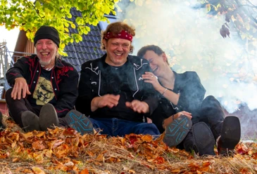 Texas Heat.png Drei Personen sitzen auf einem mit Herbstlaub bedeckten Boden vor einem Hausdach, umgeben von Rauch und Bäumen mit grünen und gelben Blättern