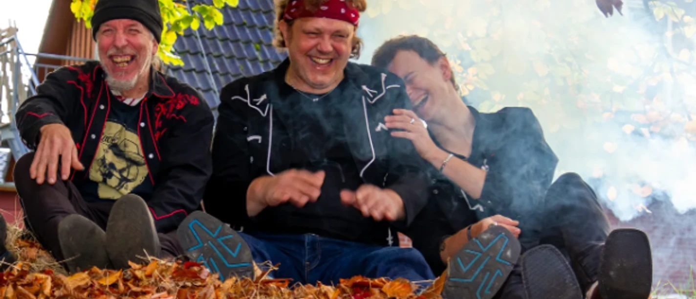 Texas Heat.png Drei Personen sitzen auf einem mit Herbstlaub bedeckten Boden vor einem Hausdach, umgeben von Rauch und Bäumen mit grünen und gelben Blättern