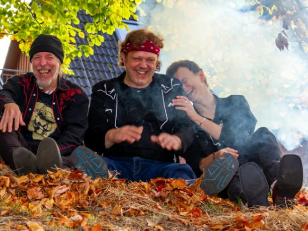 Texas Heat.png Drei Personen sitzen auf einem mit Herbstlaub bedeckten Boden vor einem Hausdach, umgeben von Rauch und Bäumen mit grünen und gelben Blättern
