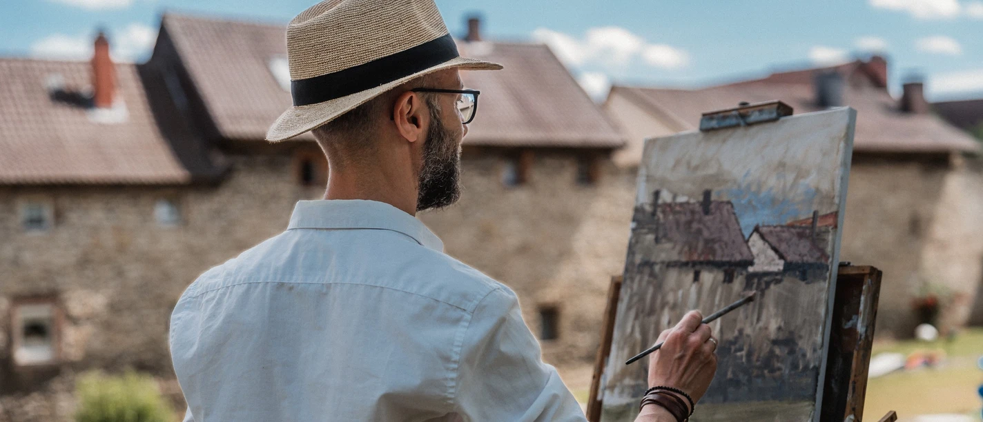 Mathias Meinel auf dem Bäckerwall, ©Timo Ehlert.jpg Ein Maler mit Strohhut malt vor historischen Gebäuden eine Landschaft auf eine Leinwand.