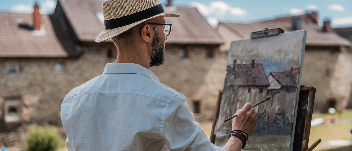 Mathias Meinel auf dem Bäckerwall, ©Timo Ehlert.jpg Ein Maler mit Strohhut malt vor historischen Gebäuden eine Landschaft auf eine Leinwand.