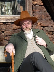 Markus Veith als Wilhelm Busch.jpg Mann in historischer Kleidung mit Pfeife und Stock sitzt lächelnd vor einem Holzhaus in der Sonne.