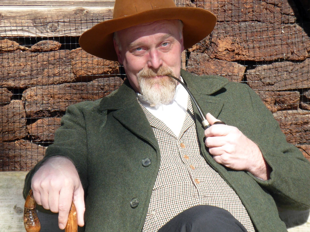 Markus Veith als Wilhelm Busch.jpg Mann in historischer Kleidung mit Pfeife und Stock sitzt lächelnd vor einem Holzhaus in der Sonne.