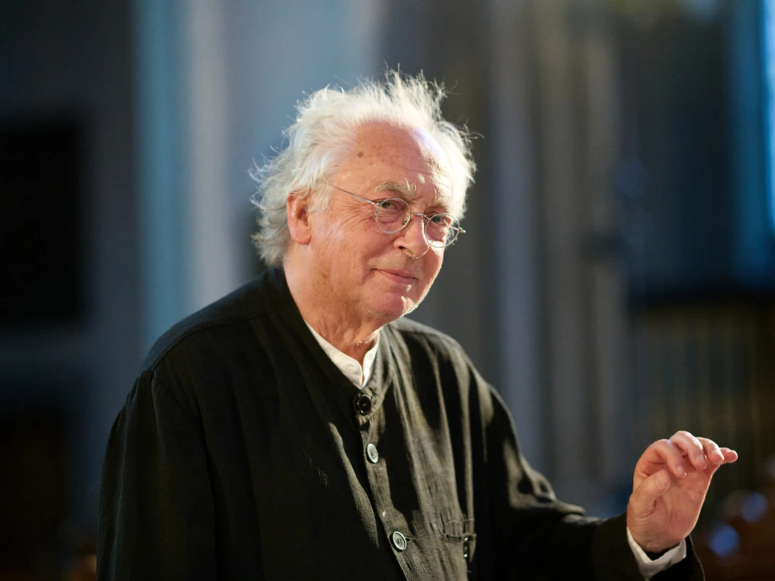 Philippe Herreweghe Älterer Mann mit Brille in dunkler Kleidung hebt die Hand in einem lichtdurchfluteten Raum.