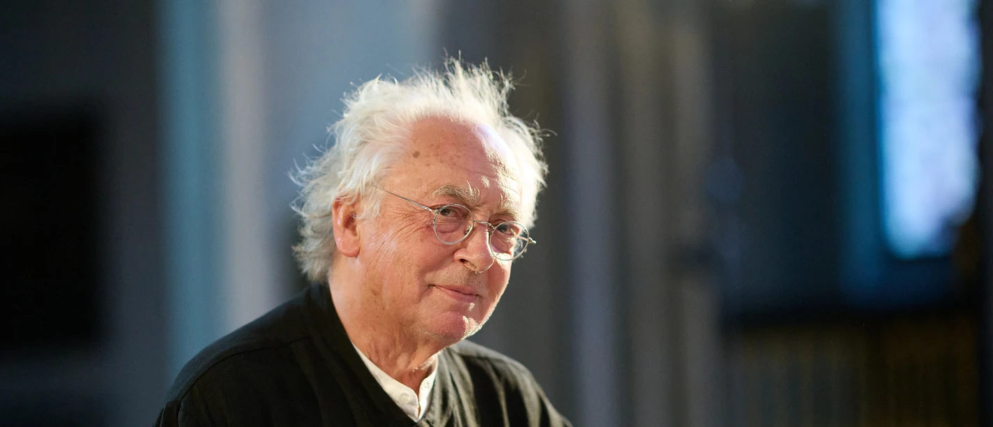 Philippe Herreweghe Älterer Mann mit Brille in dunkler Kleidung hebt die Hand in einem lichtdurchfluteten Raum.
