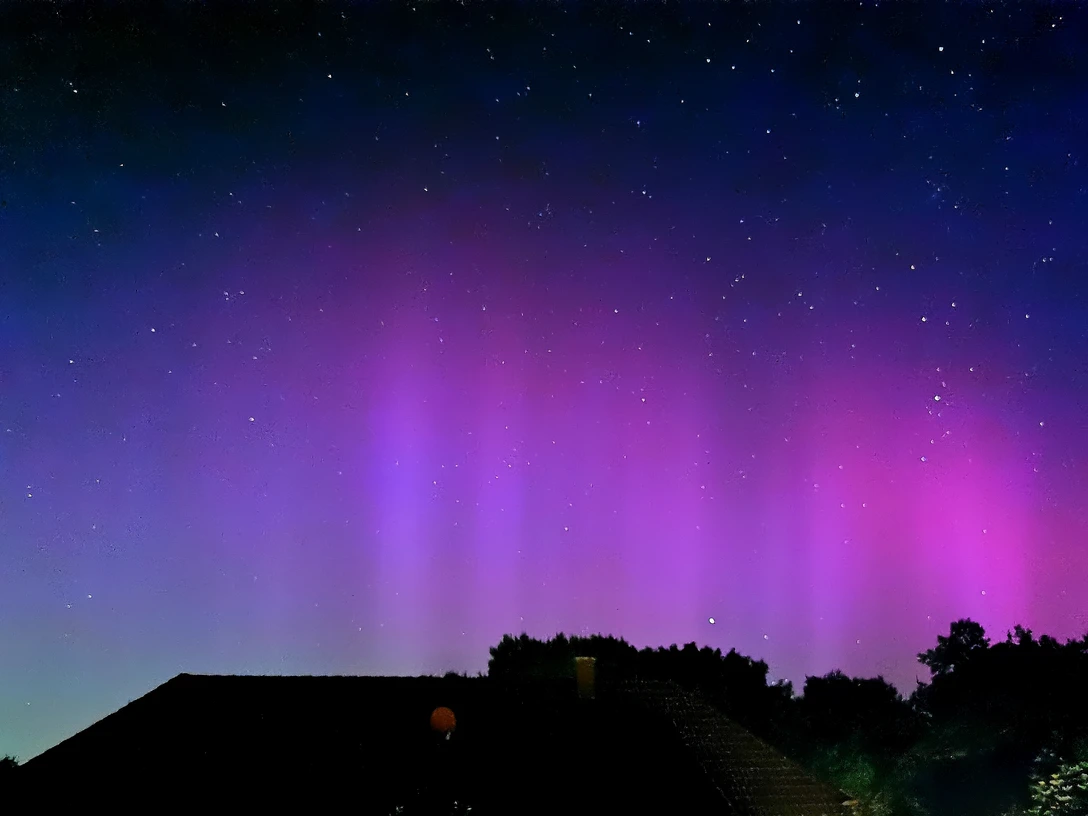 Polarlichter 2.1.jpg Am Nachthimmel leuchten violette und rosafarbene Polarlichter über dunklen Dächern und Baumkronen, während zahlreiche Sterne in der klaren Himmelskuppe funkeln.