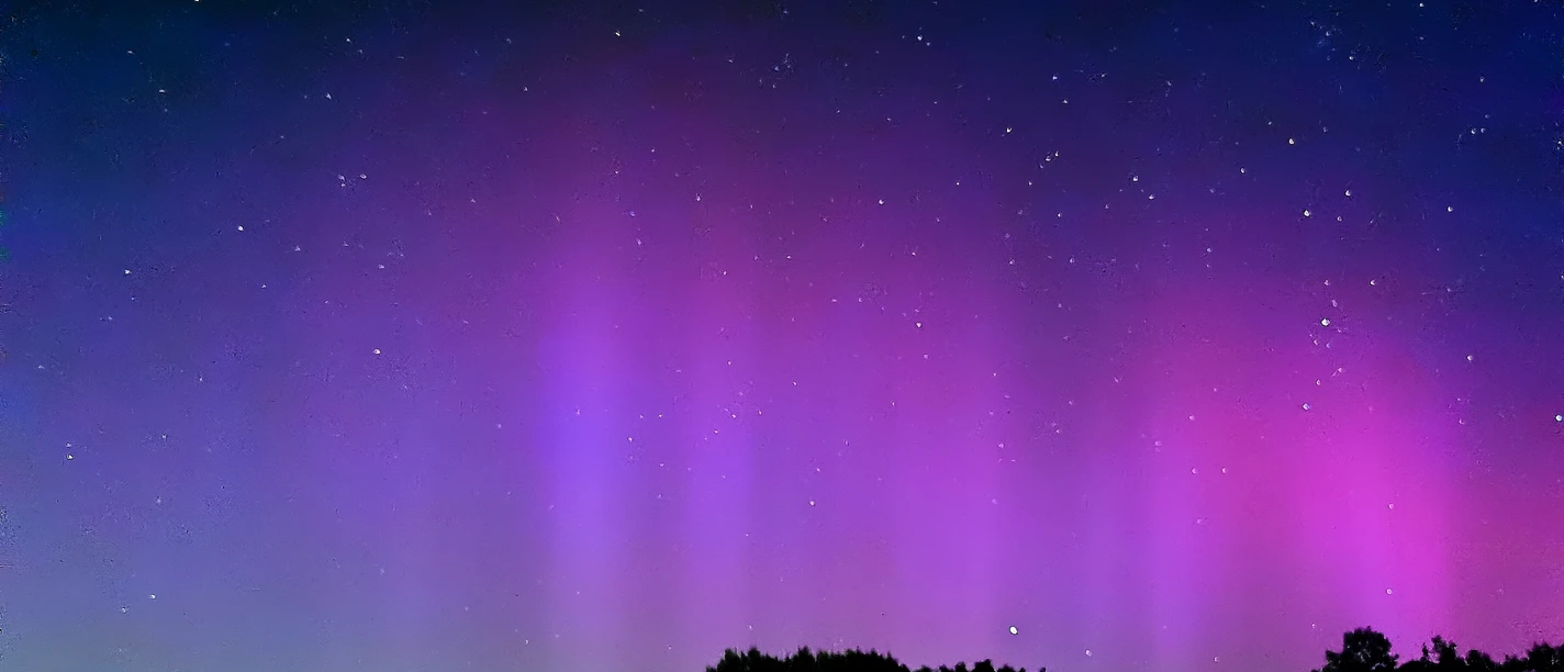 Polarlichter 2.1.jpg Am Nachthimmel leuchten violette und rosafarbene Polarlichter über dunklen Dächern und Baumkronen, während zahlreiche Sterne in der klaren Himmelskuppe funkeln.