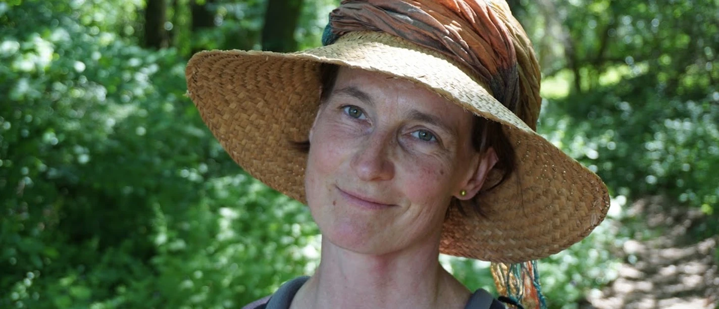 Kräuterspaziergang mit Doerte Fähling Frau mit Strohhut und Rucksack lächelt auf einem Waldweg, umgeben von grünem Laub.