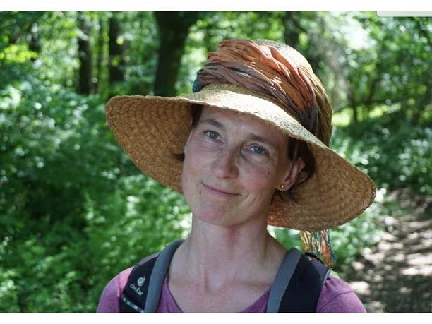 Kräuterspaziergang mit Doerte Fähling Frau mit Strohhut und Rucksack lächelt auf einem Waldweg, umgeben von grünem Laub.