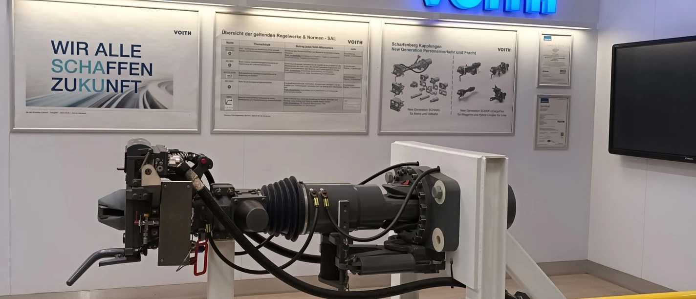 2026_Betriebsbesichtigung_Voith (1)_HD_16zu9.jpg Ausstellungsraum bei Voith mit beleuchtetem Firmenlogo und Exponat einer modernen Zugkupplung.