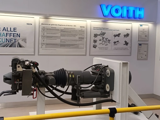 2026_Betriebsbesichtigung_Voith (1)_HD_16zu9.jpg Ausstellungsraum bei Voith mit beleuchtetem Firmenlogo und Exponat einer modernen Zugkupplung.